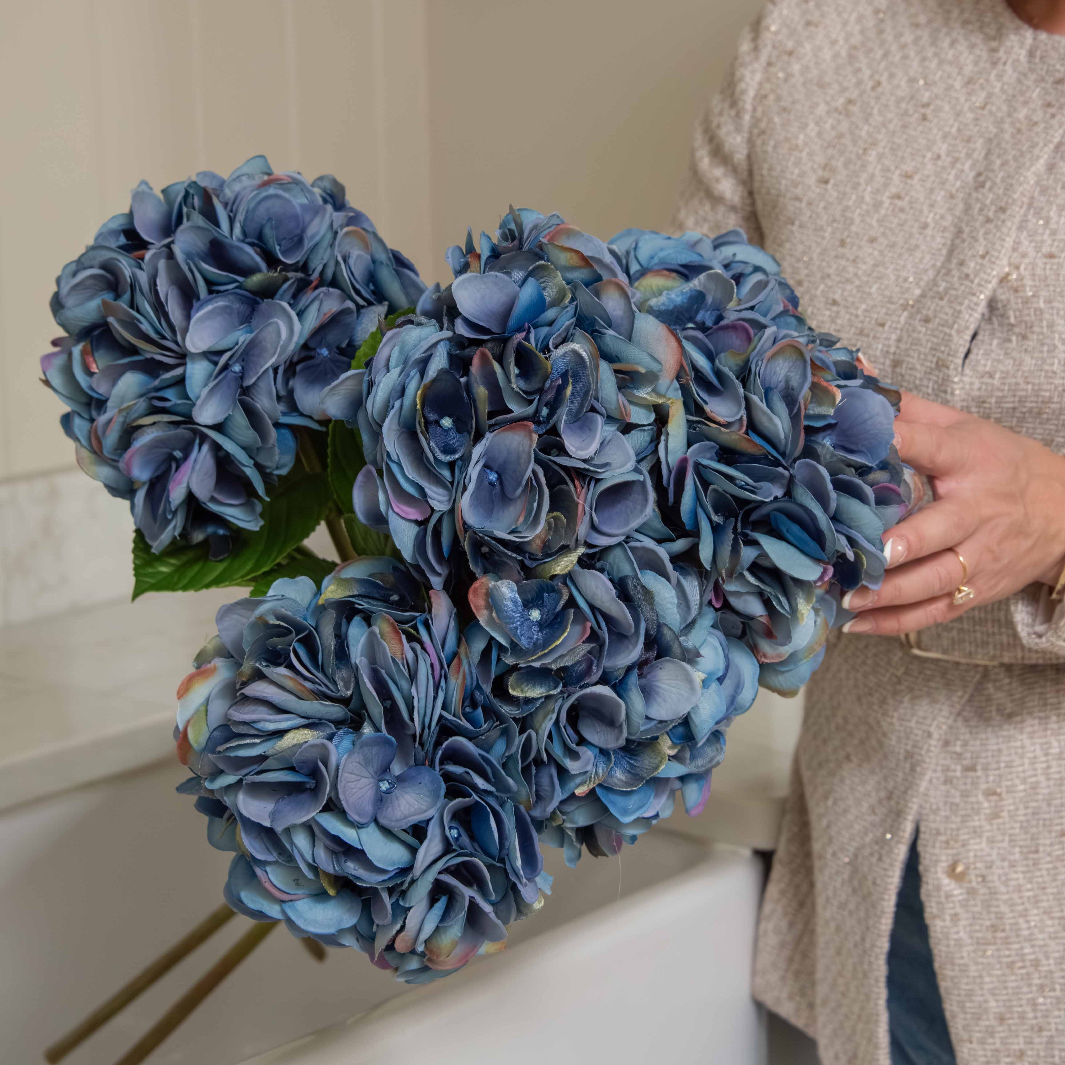 A person holding a faux blue hydrangea stem with realistic petals and leaves.