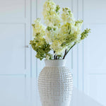A single lilac stem in a white vase, with green and white leaves, placed on a white surface against a white background.