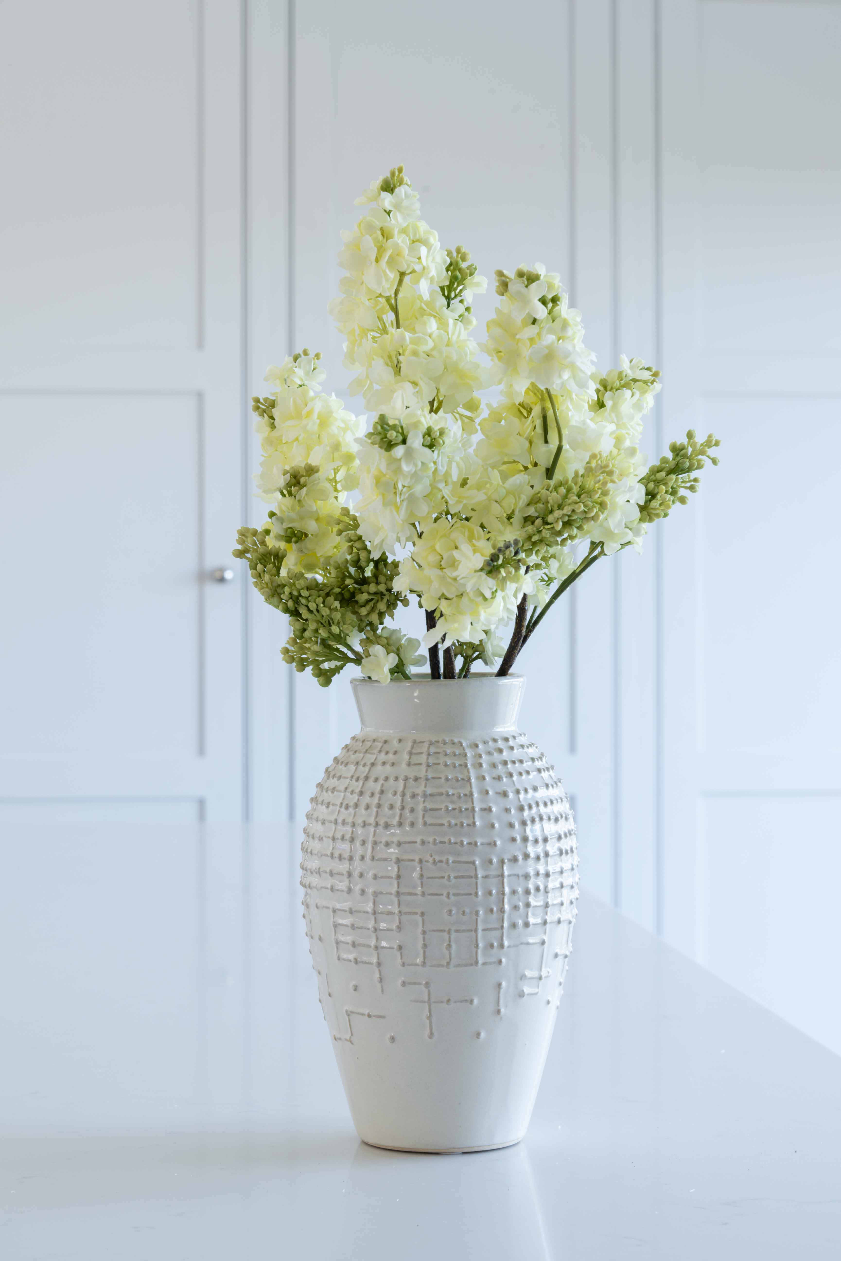 A single lilac stem in a white vase, with green and white leaves, placed on a white surface against a white background.