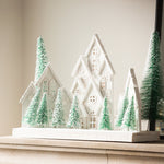 Charles Ted White Christmas Village Scene with snowy white houses and frosted green trees displayed on a mantel