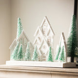 Charles Ted White Christmas Village Scene with snowy white houses and frosted green trees displayed on a mantel