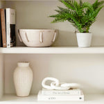 A stunning white ceramic vase sitting on a bookshelf. There is a raised-bump pattern on the exterior of the vase