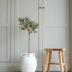 A faux olive tree with green leaves in a white ceramic garden pot, placed on a floor next to a wooden stool, with a closed white door in the background.