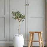 A faux olive tree with green leaves in a white ceramic garden pot, placed on a floor next to a wooden stool, with a closed white door in the background.