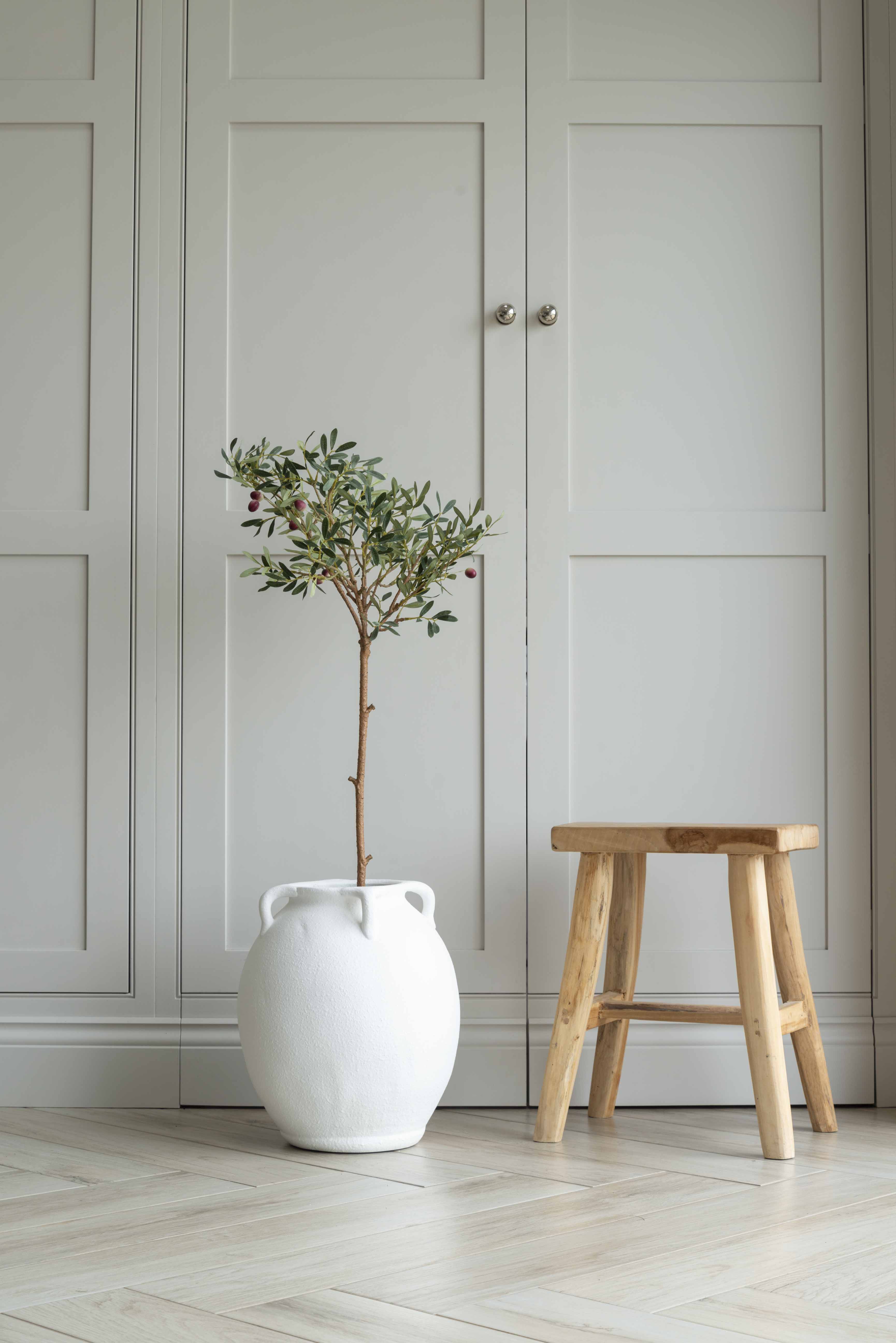 A faux olive tree with green leaves in a white ceramic garden pot, placed on a floor next to a wooden stool, with a closed white door in the background.