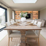 A rustic wooden dining table with a central pedestal base, surrounded by four chairs, set in a well-lit room with a large window.