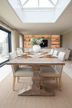 A rustic wooden dining table with a central pedestal base, surrounded by four chairs, set in a well-lit room with a large window.