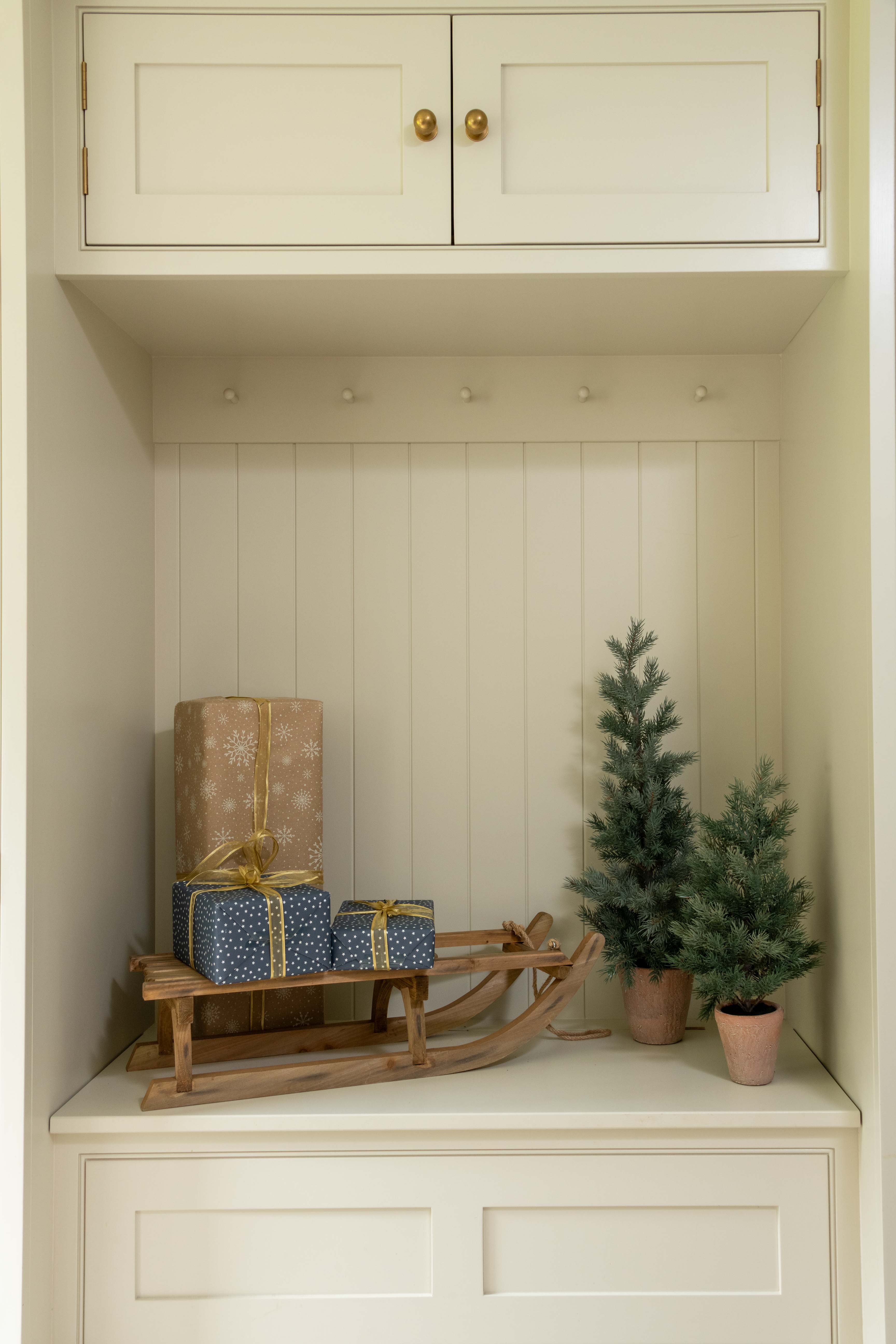Nook with wooden sled, wrapped presents, and small Christmas trees inside a white cabinet.