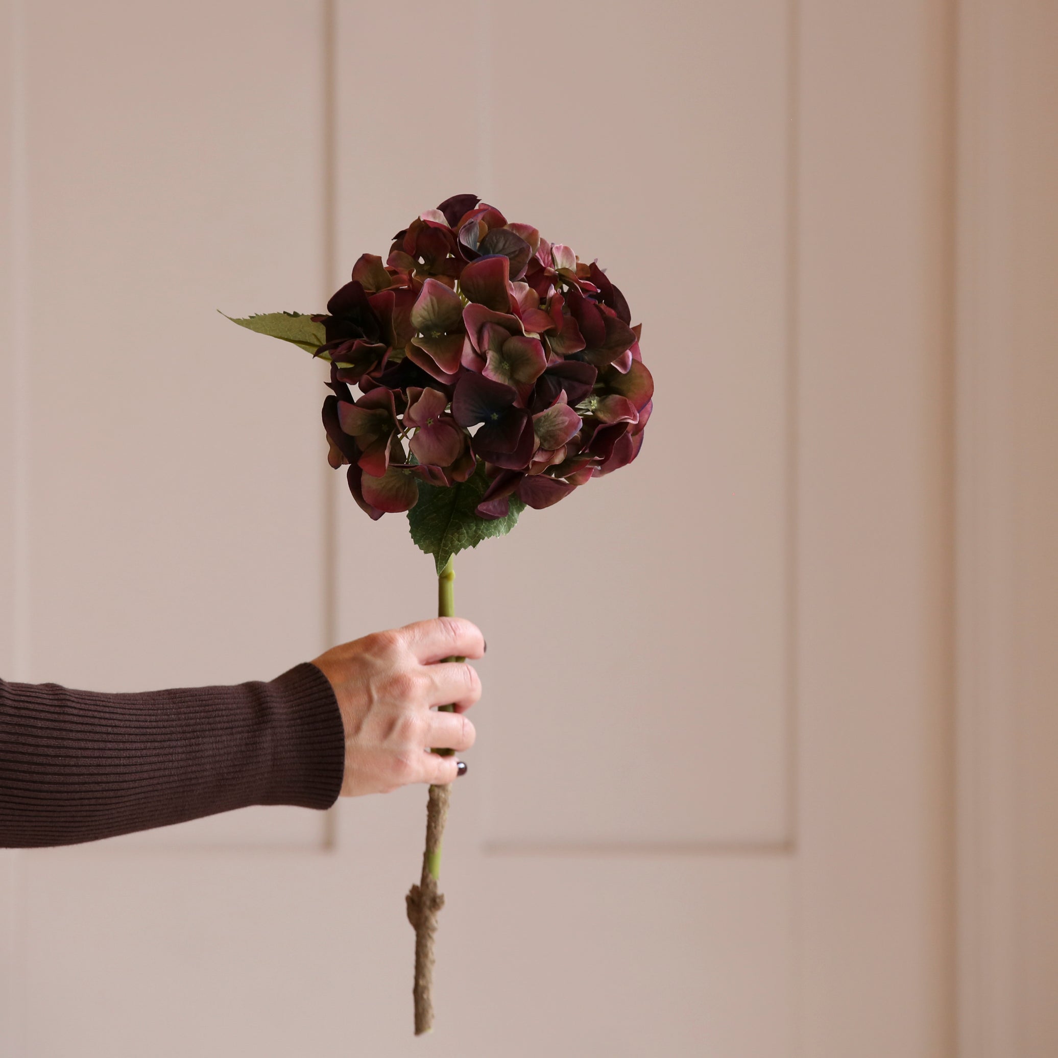 A person holding a lifelike faux burgundy hydrangea flower with hints of green.