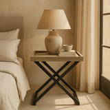 A rectangular oak side table with black metal legs in a bedroom setting, featuring a lamp and some books on top.