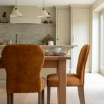 A burnt orange velvet dining chair with brass stud detailing, pictured in a dining setting with a wooden table and pendant lights overhead.