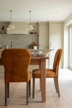A burnt orange velvet dining chair with brass stud detailing, pictured in a dining setting with a wooden table and pendant lights overhead.