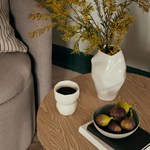 White vase with yellow flowers on a wooden table next to a cup of coffee and a bowl of figs.