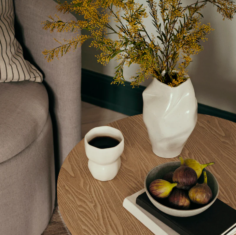 White vase with yellow flowers on a wooden table next to a cup of coffee and a bowl of figs.