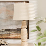 A white marble table lamp with a linen shade placed on a wooden table next to a framed picture on the wall, with a green plant in the foreground.