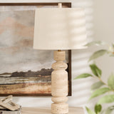 A white marble table lamp with a linen shade placed on a wooden table next to a framed picture on the wall, with a green plant in the foreground.