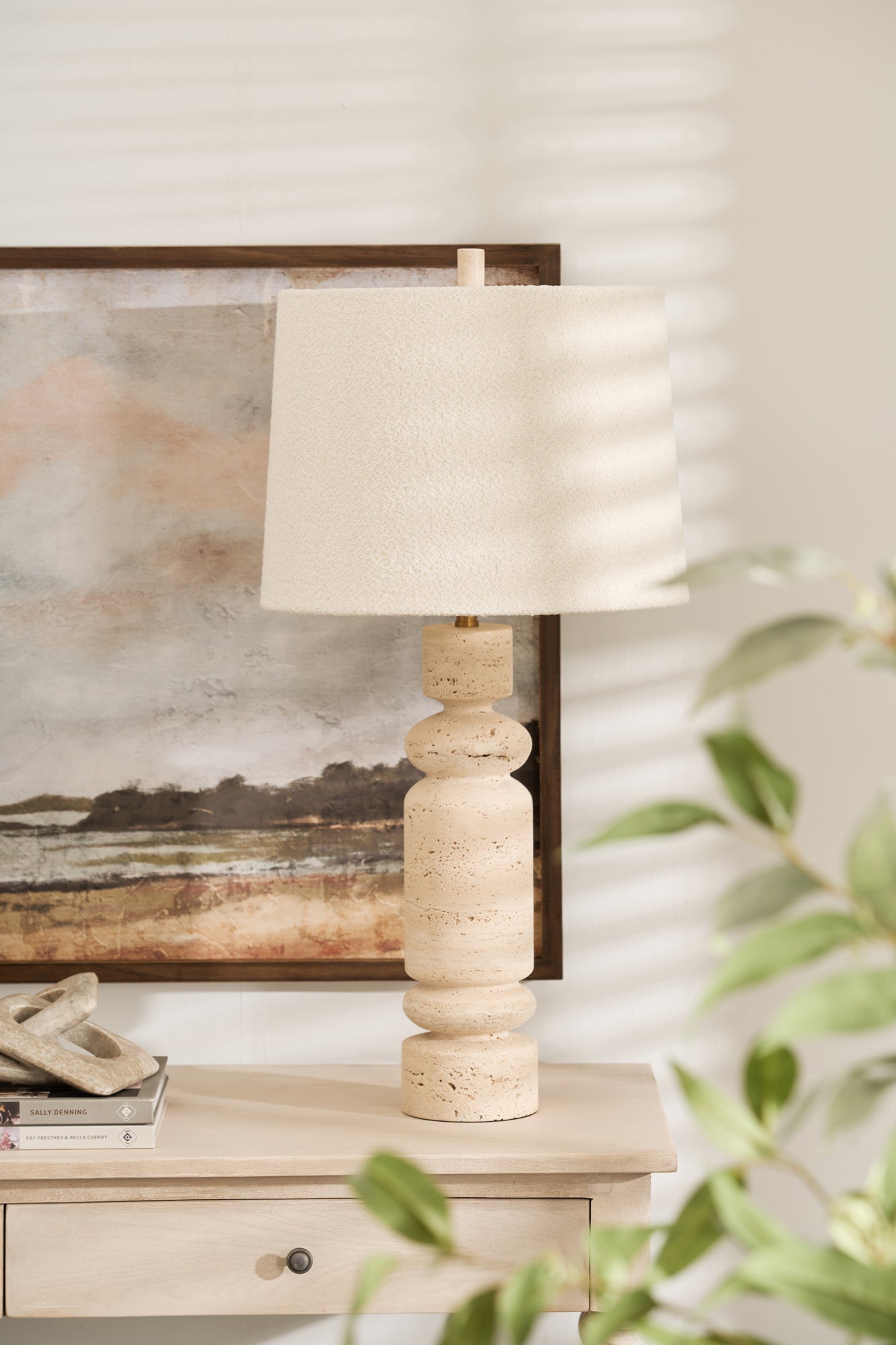 A white marble table lamp with a linen shade placed on a wooden table next to a framed picture on the wall, with a green plant in the foreground.