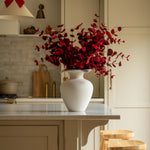 Kitchen with a white vase of red flowers on a counter, wooden stools, and a red bow decoration.