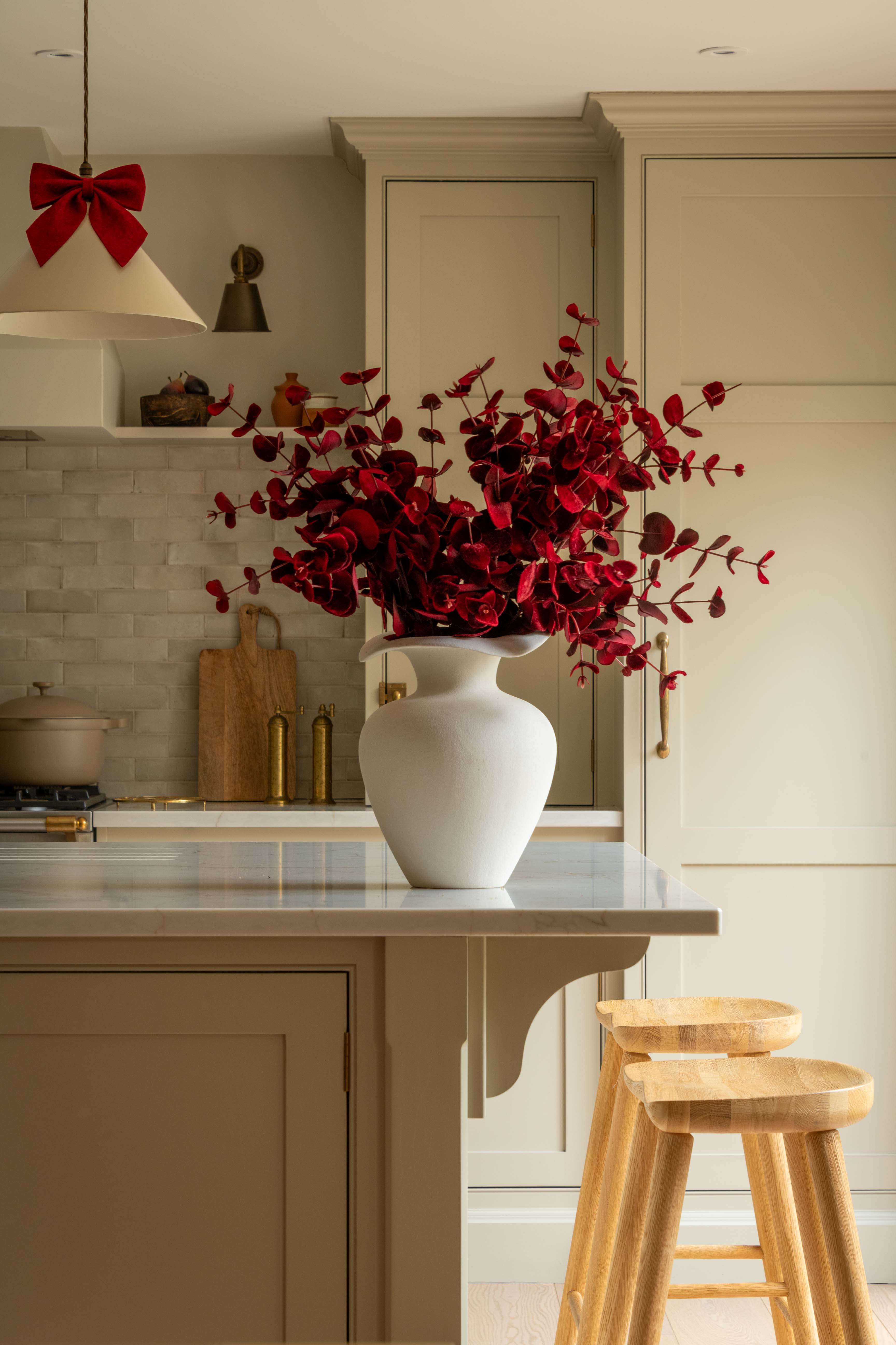 Kitchen with a white vase of red flowers on a counter, wooden stools, and a red bow decoration.