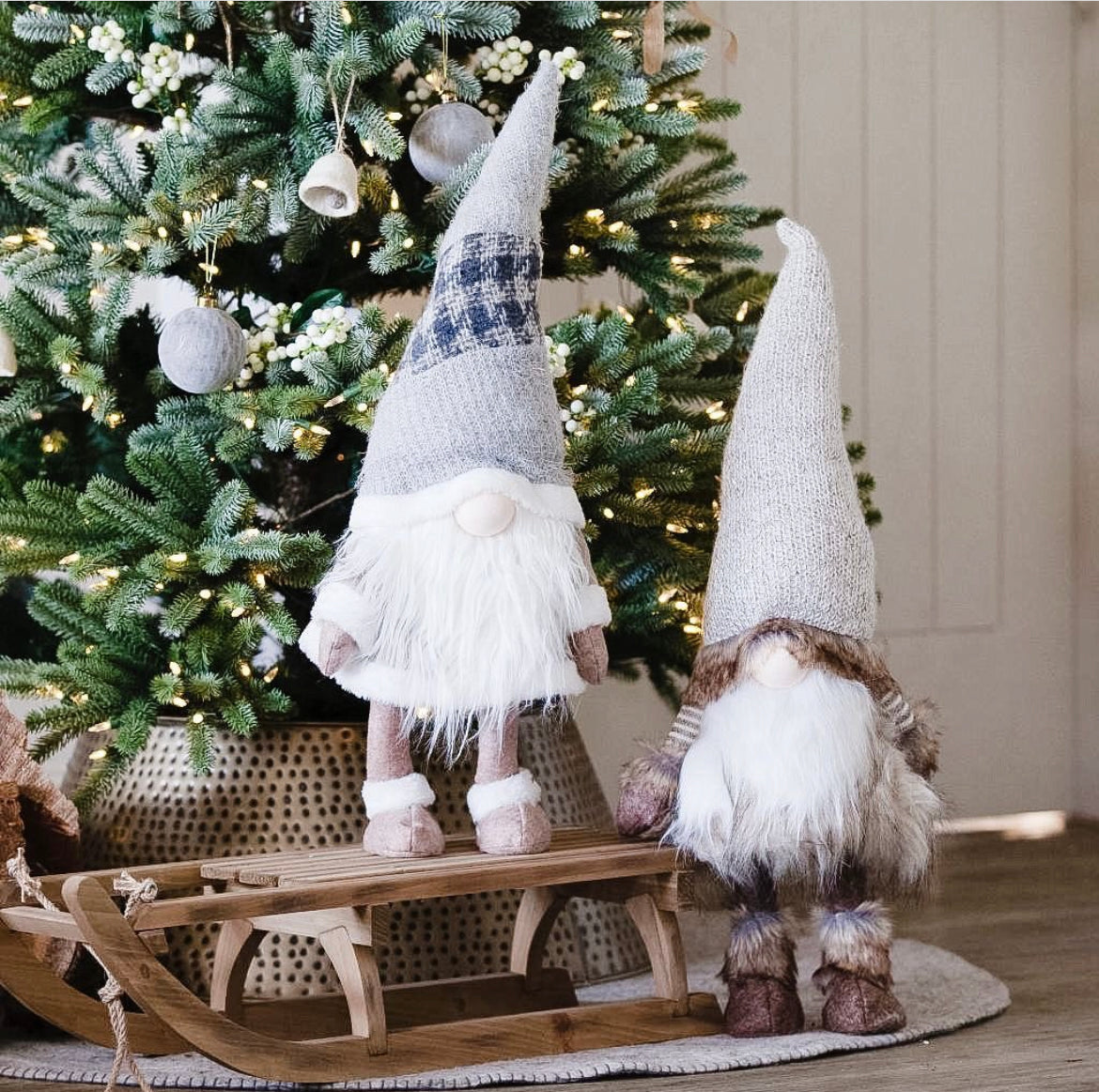 Two figurines of bearded elves stand on a wooden sleigh, under a Christmas tree