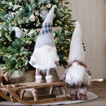 Two figurines of bearded elves stand on a wooden sleigh, under a Christmas tree
