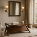 An acacia wood coffee table with a glass top, featuring a sleek design, placed in a room with a mirror on the wall, a candle, and a book on top.