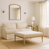 Bright living room with white shiplap wall paneling, a large mirror flanked by brass wall sconces, a beige linen sofa, a wooden side table with a white lamp, long linen curtains over a large window, and a square light wood coffee table with a cushioned top on a textured rug.