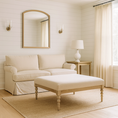 Bright living room with white shiplap wall paneling, a large mirror flanked by brass wall sconces, a beige linen sofa, a wooden side table with a white lamp, long linen curtains over a large window, and a square light wood coffee table with a cushioned top on a textured rug.