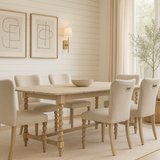 A bright dining room with white shiplap walls, a light wood dining table with spindle legs, and  matching beige upholstered chairs. The set sits on a textured jute rug over light wood floors. Above the table hangs abstract art flanked by two black sconces with white shades. A large window with beige linen curtains lets in natural light, and an olive tree in a woven basket stands in the corner