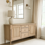 A light oak sideboard with panelled doors and three central drawers sits against a white shiplap wall in a bright living room. Above it hangs a large rectangular mirror with a gold frame, flanked by two brass wall sconces with white shades. A neutral ceramic table lamp with a fabric shade rests on the left side of the sideboard. Beige linen curtains hang beside a large window, letting in soft natural light, and a light textured rug covers the wooden floor.