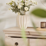 A faux ranunculus flower arrangement in a cream-colored vase, displayed on a wooden surface with a drawer.