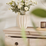 A faux ranunculus flower arrangement in a cream-colored vase, displayed on a wooden surface with a drawer.
