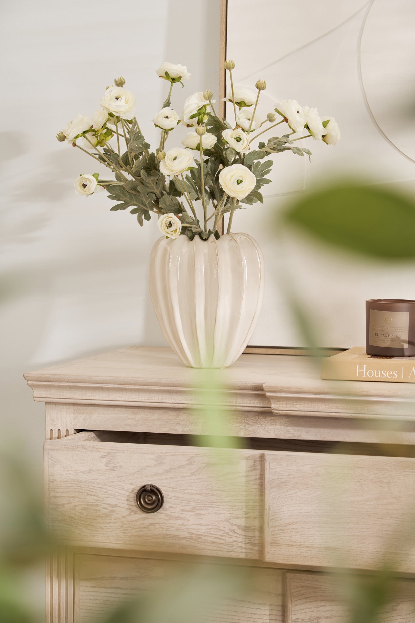 A faux ranunculus flower arrangement in a cream-colored vase, displayed on a wooden surface with a drawer.