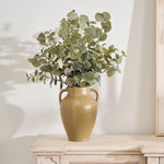 An olive green handled urn with rustic finish handles, filled with greenery, placed on a wooden surface.