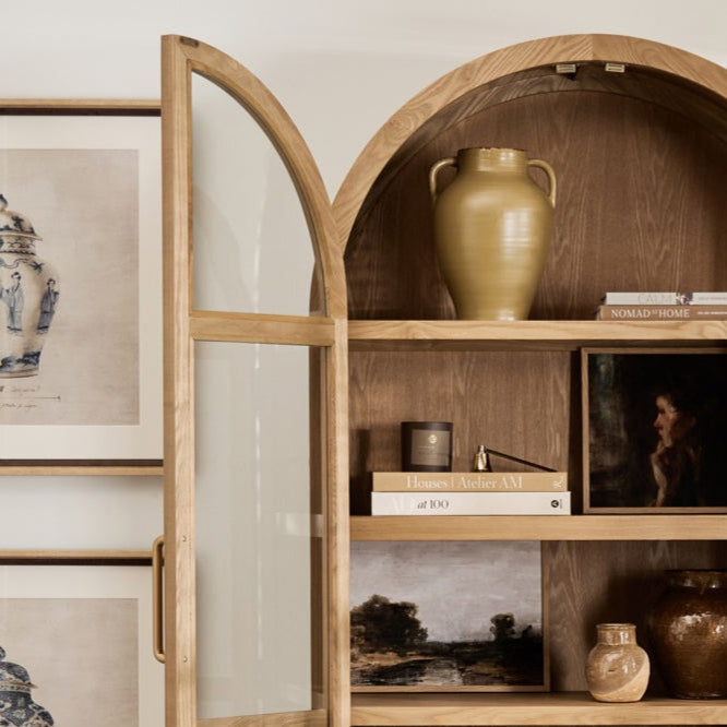Wooden armoire with open doors displaying various items in a room with framed pictures and a plant.