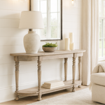 A rustic wooden console table with turned legs is placed against a white shiplap wall in a bright living room. Above the table hangs a large, black-framed arched mirror, flanked by two brass wall sconces with glass shades. On the table are a ceramic lamp with a white shade, a small bowl of greenery, and several pillar candles. Light linen curtains frame a large window, allowing natural light to fill the space.