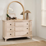 A wooden arched mirror mounted above a dresser with multiple drawers. The room it's placed in appears to be well-lit with natural light and has a window in the background.
