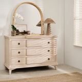 A wooden arched mirror mounted above a dresser with multiple drawers. The room it's placed in appears to be well-lit with natural light and has a window in the background.