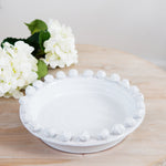 A white ceramic fruit bowl with a decorative edge, displayed on a wooden surface with white flowers in the background.