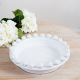 A white ceramic fruit bowl with a decorative edge, displayed on a wooden surface with white flowers in the background.