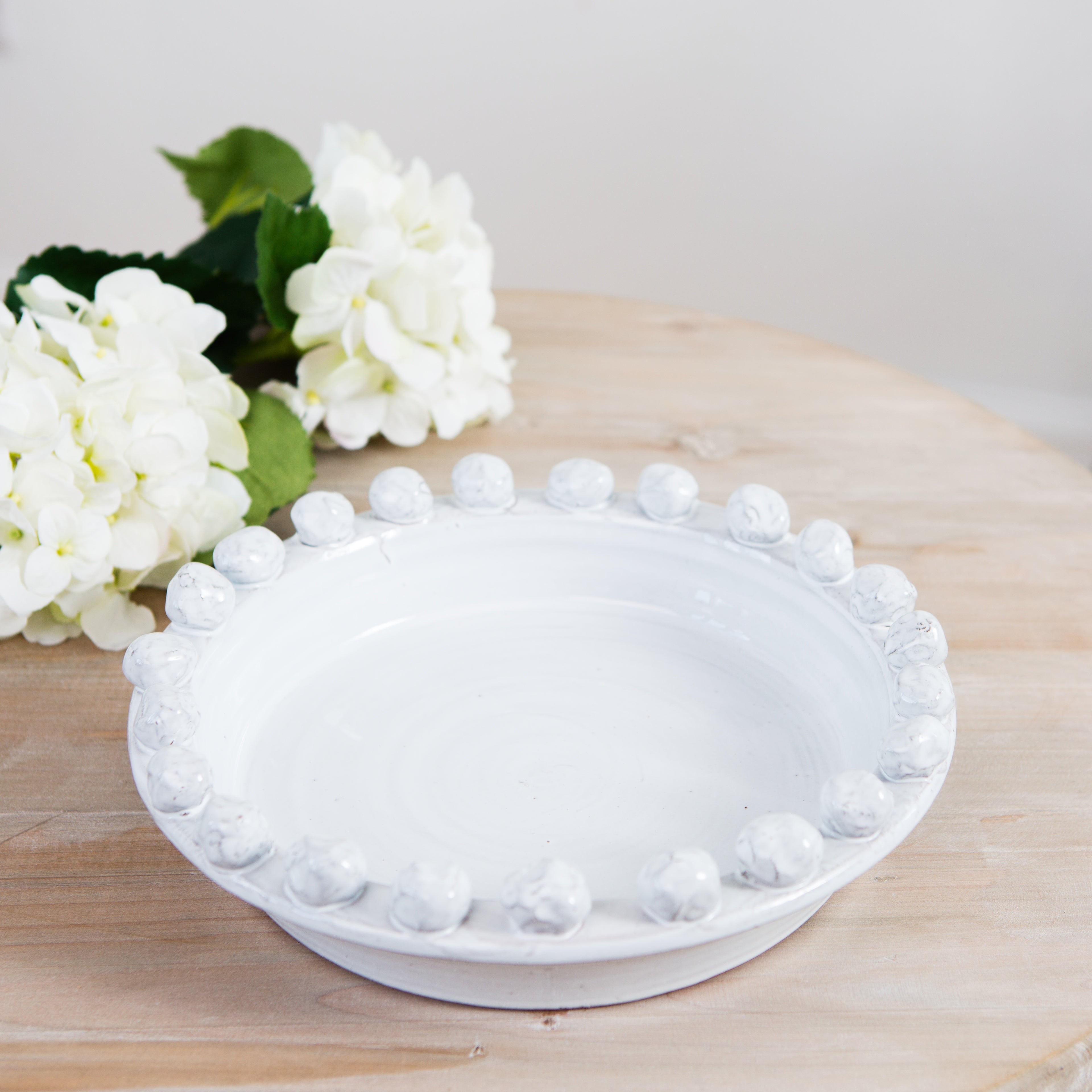 A white ceramic fruit bowl with a decorative edge, displayed on a wooden surface with white flowers in the background.
