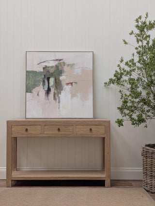 A light oak wood console table with three drawers and a lower shelf, styled in a room setting with an abstract painting on the wall and a potted green plant.