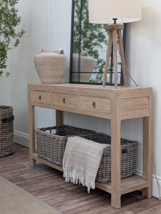 A light oak wood console table with three drawers and an open lower shelf, styled in a home entryway with a woven storage basket, a tripod floor lamp, and a mirror.