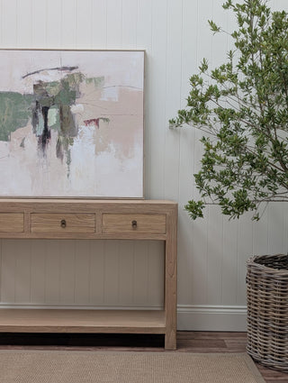 A light natural wood console table with three drawers and a lower shelf, styled in a room setting with an abstract painting on the wall and a potted green plant in a wicker basket.