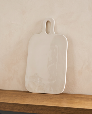 A white ceramic serving board with a rounded handle, resting on a wooden shelf against a textured beige wall.