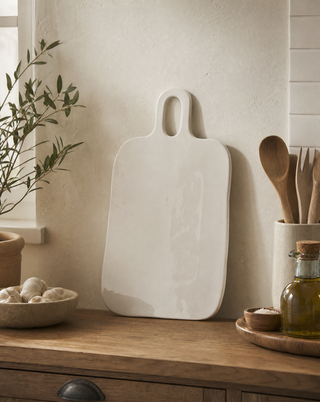 A white ceramic serving board with a loop handle, leaning on a wooden kitchen counter next to a bowl of garlic, a plant, and wooden utensils.