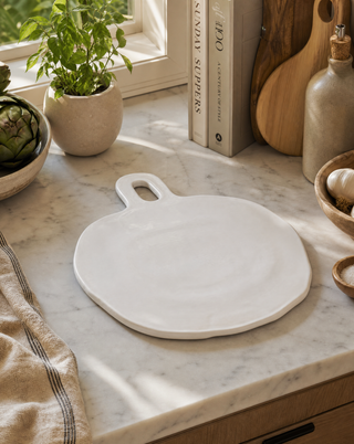 A round white ceramic serving board with a handle, placed on a white marble kitchen countertop surrounded by home decor items like a potted plant, books, and wooden cutting boards.