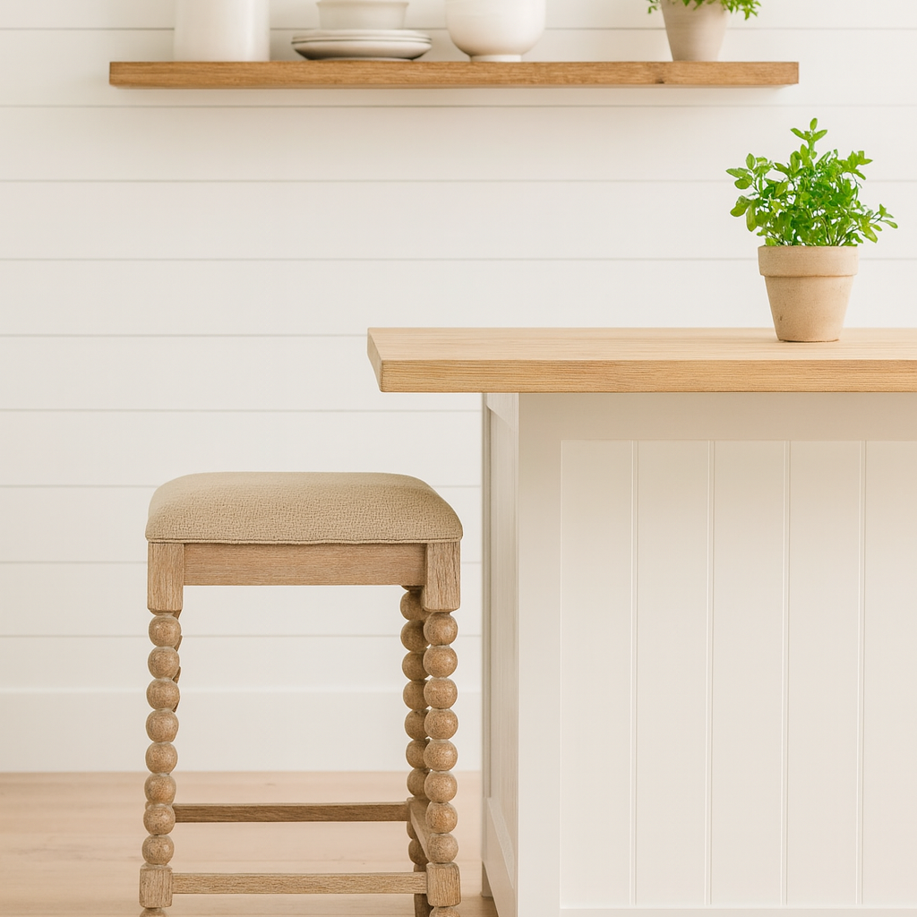 A farmhouse-style kitchen with white shiplap walls, a wooden floating shelf holding a white ceramic pitcher, stacked plates, bowls, and a small potted herb plant. Below, a light wood and white kitchen island with a beige-upholstered stool featuring turned wooden legs sits on a light hardwood floor, accompanied by another potted herb on the island countertop.