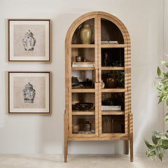 Wooden arched cabinet with glass doors in a room with framed pictures and a plant.