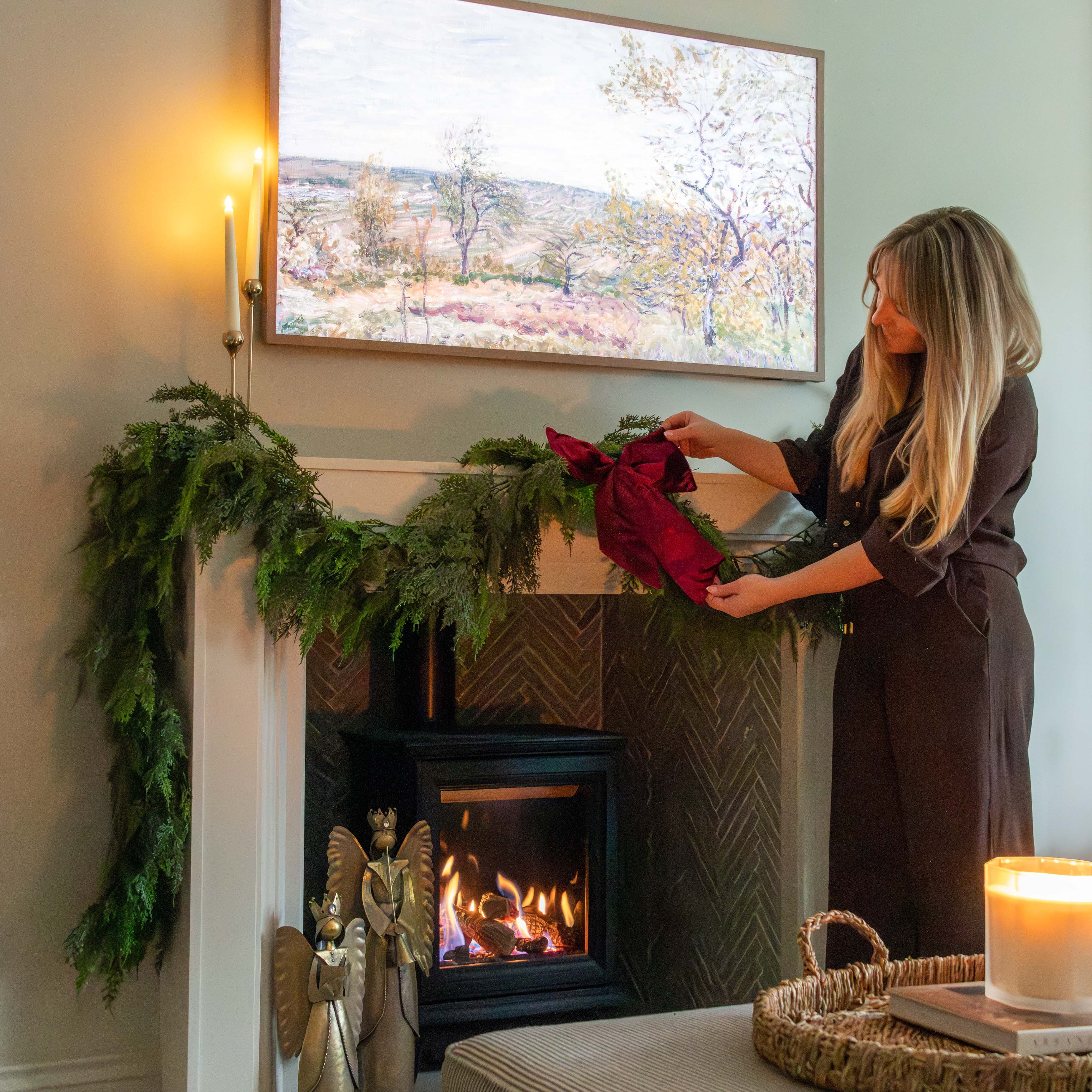 Woman decorating a fireplace with greenery and a red bow in a cozy living room.